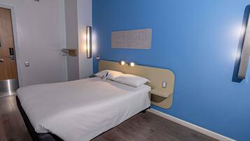 Una habitación de hotel con una cama, una lámpara fijada a la pared, una puerta y una pared azul.