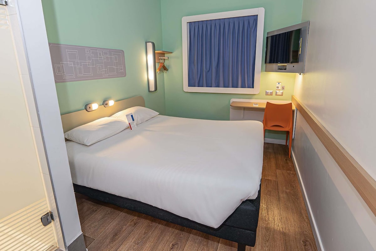A hotel room with a bed, bedside table, chair, and a TV mounted on the wall.