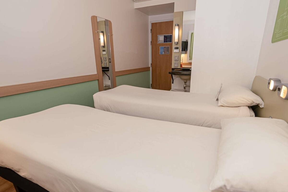 Habitación de hotel con dos camas individuales, cabecero de madera, un espejo, baño pequeño con lavabo y pared verde.