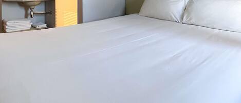 Una habitación de hotel con una cama grande, una mesita de noche con lámpara y un baño a la vista en el fondo.