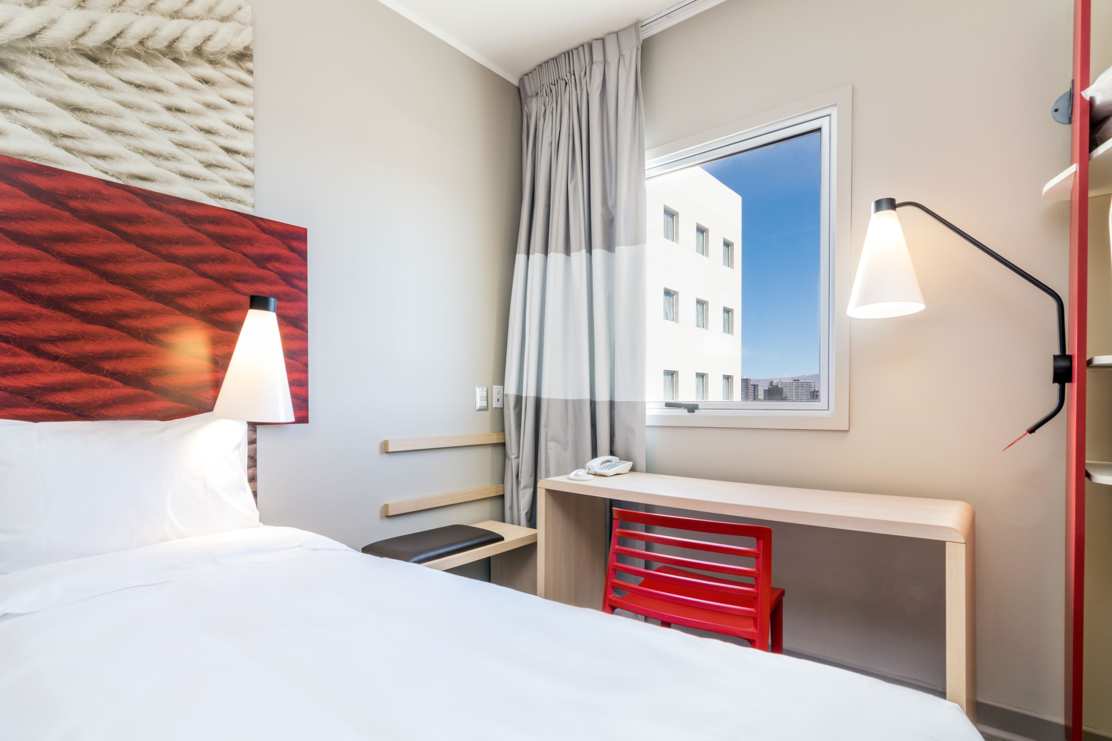 Une chambre d’hôtel comprenant un lit, un bureau, une chaise, une lampe et une fenêtre donnant sur un immeuble.