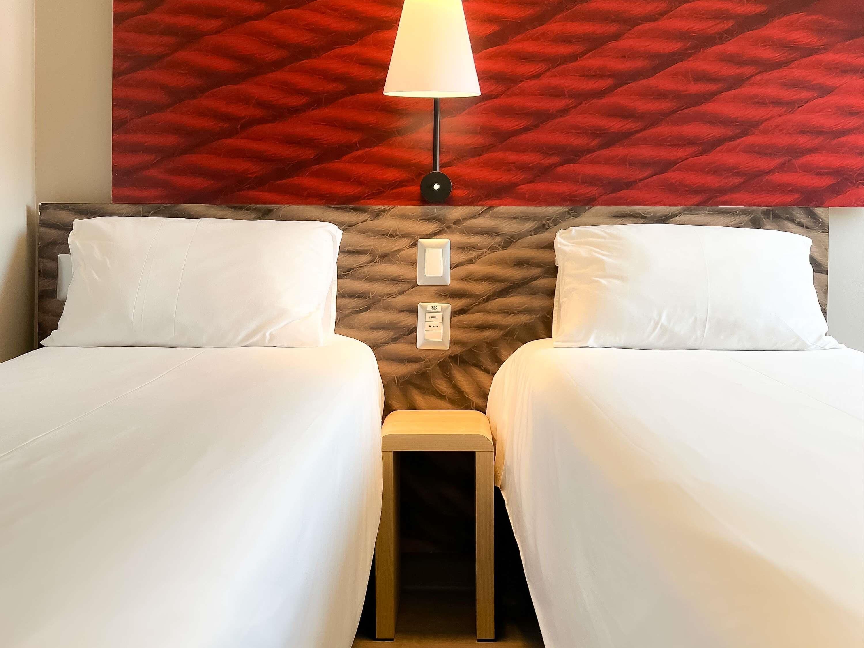 Una habitación de hotel con dos camas, una pared roja con textura, una mesita de noche y una lámpara.