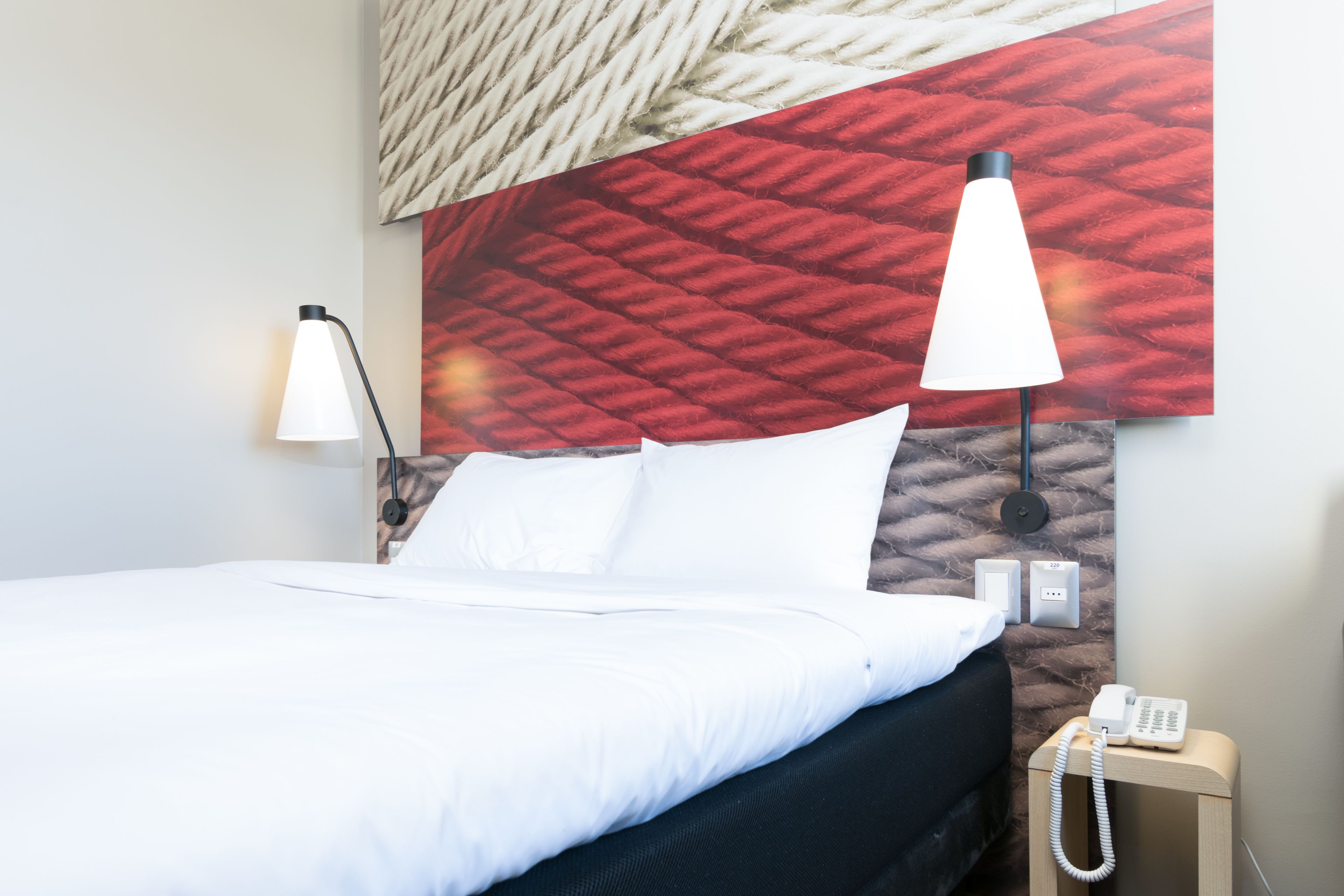 Hotellihuone, jossa on sänky, kaksi yöpöytälamppua, puhelin ja sängyn takana kuvioitu seinä.