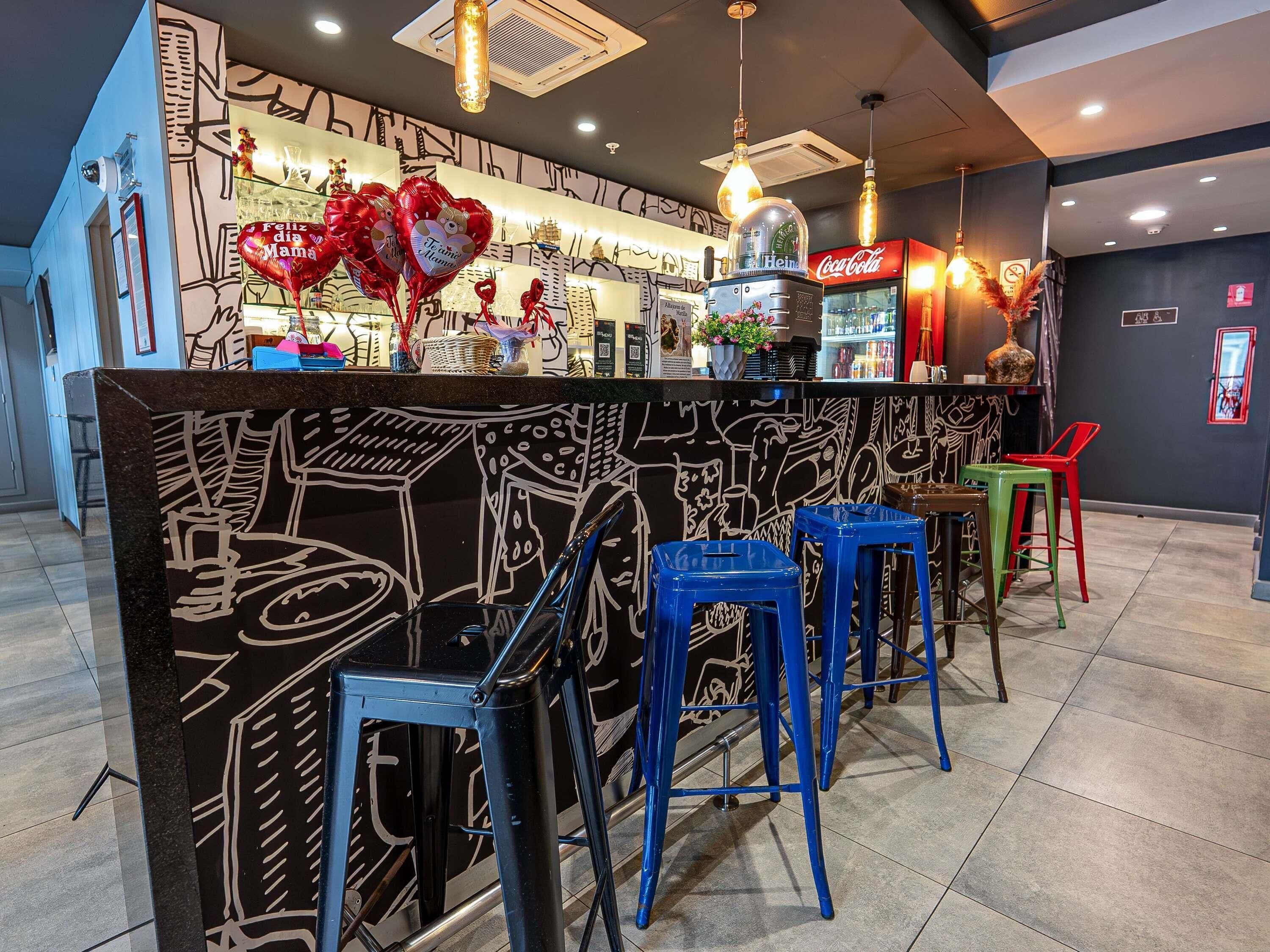 Zona de bar con un mural en blanco y negro, taburetes azules y rojos, y un refrigerador de Coca-Cola.