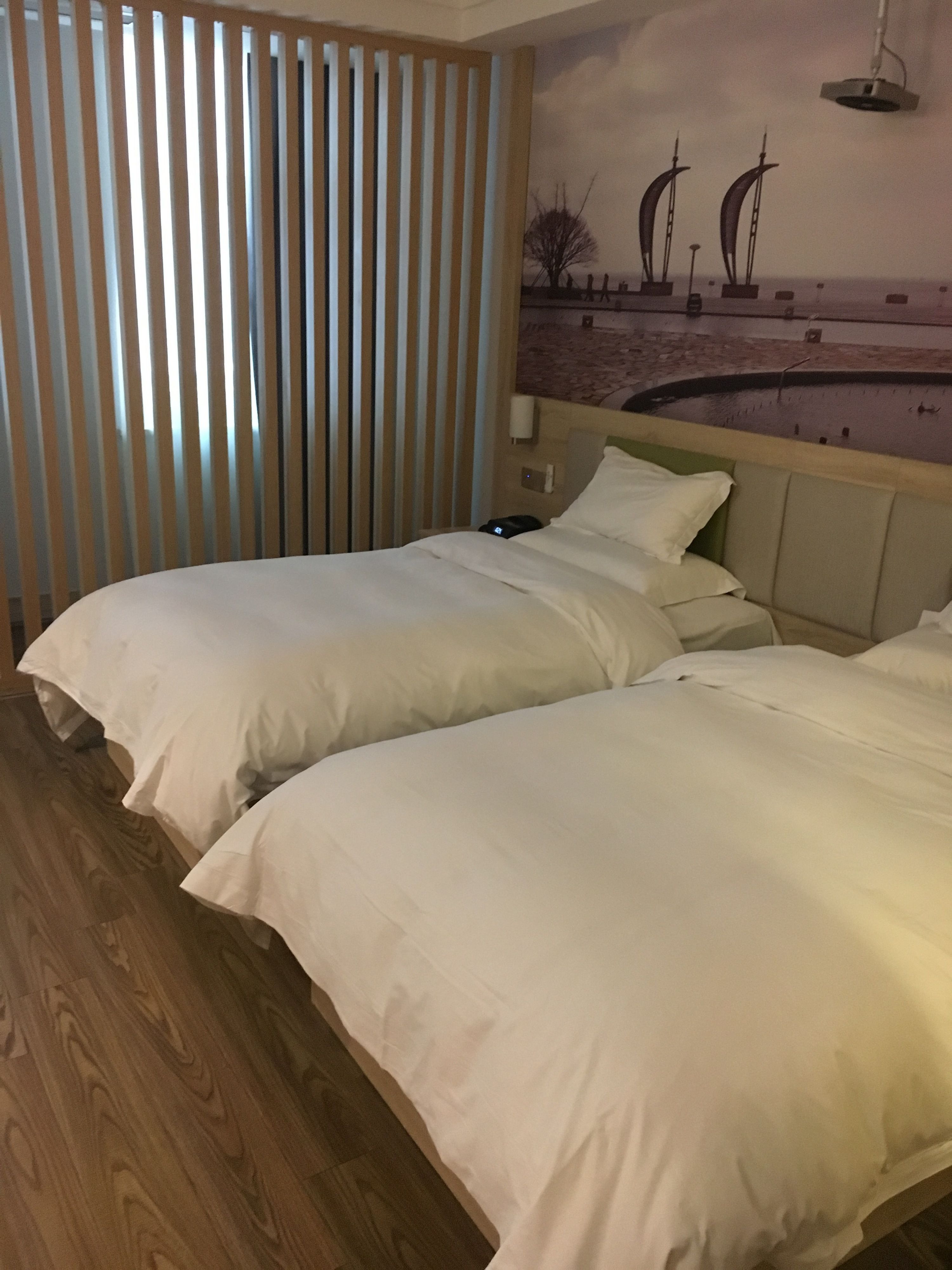 A hotel room with two beds, wooden flooring, a wall-mounted lamp, and a mural of sailboats.