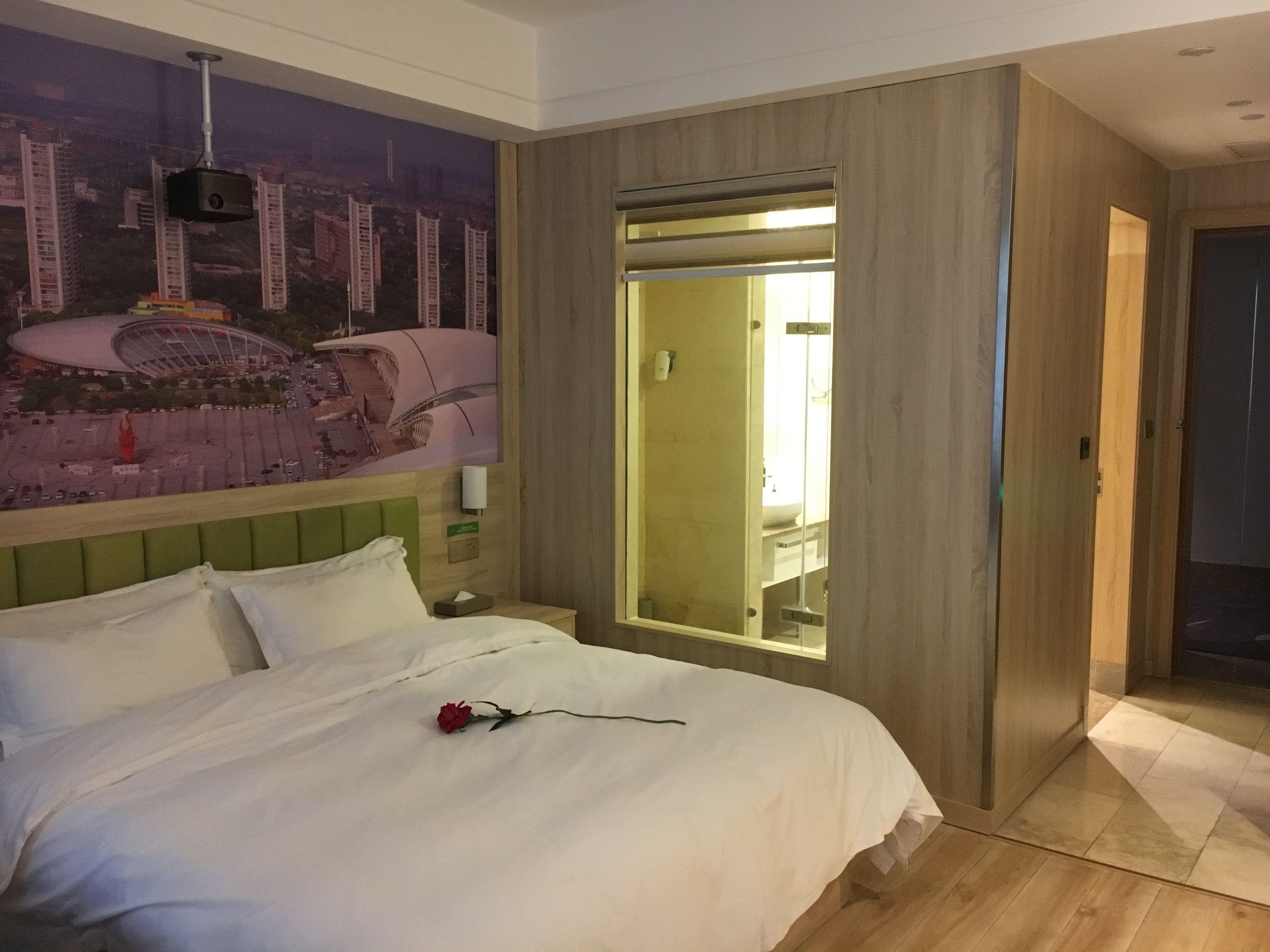 A hotel room with a bed, a green headboard, a wall-mounted projector, a large window with a view of buildings, and a wooden wardrobe.