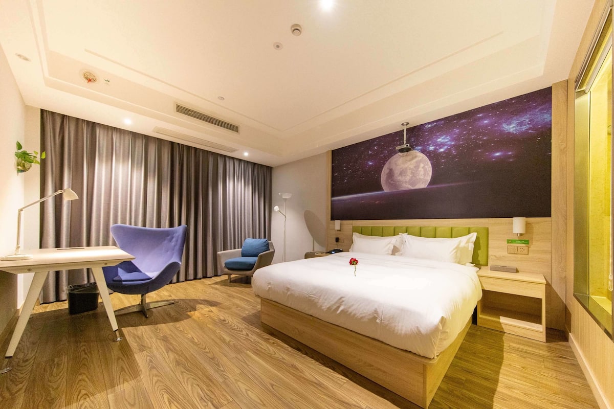 A modern hotel room with a large bed, a desk, a chair, and a wall mural of a celestial scene.