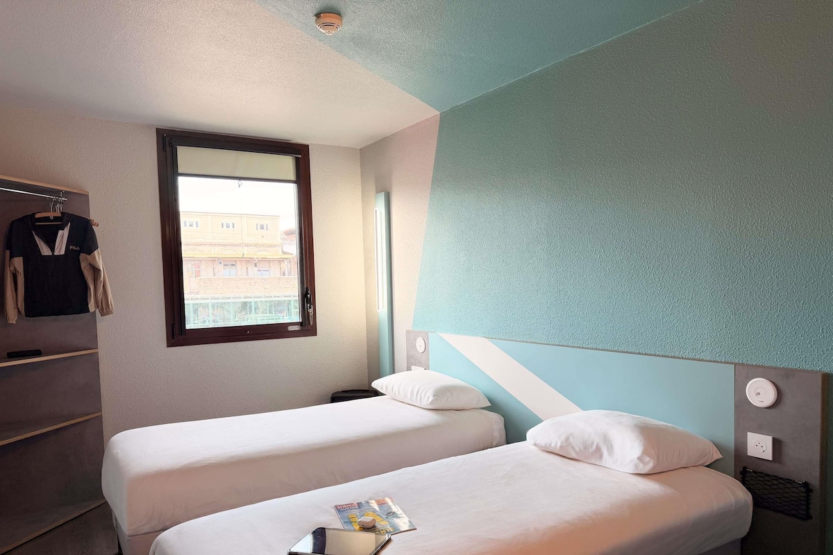 Une chambre d’hôtel avec deux lits, une fenêtre, un radiateur fixé au mur et un porte-vêtement.