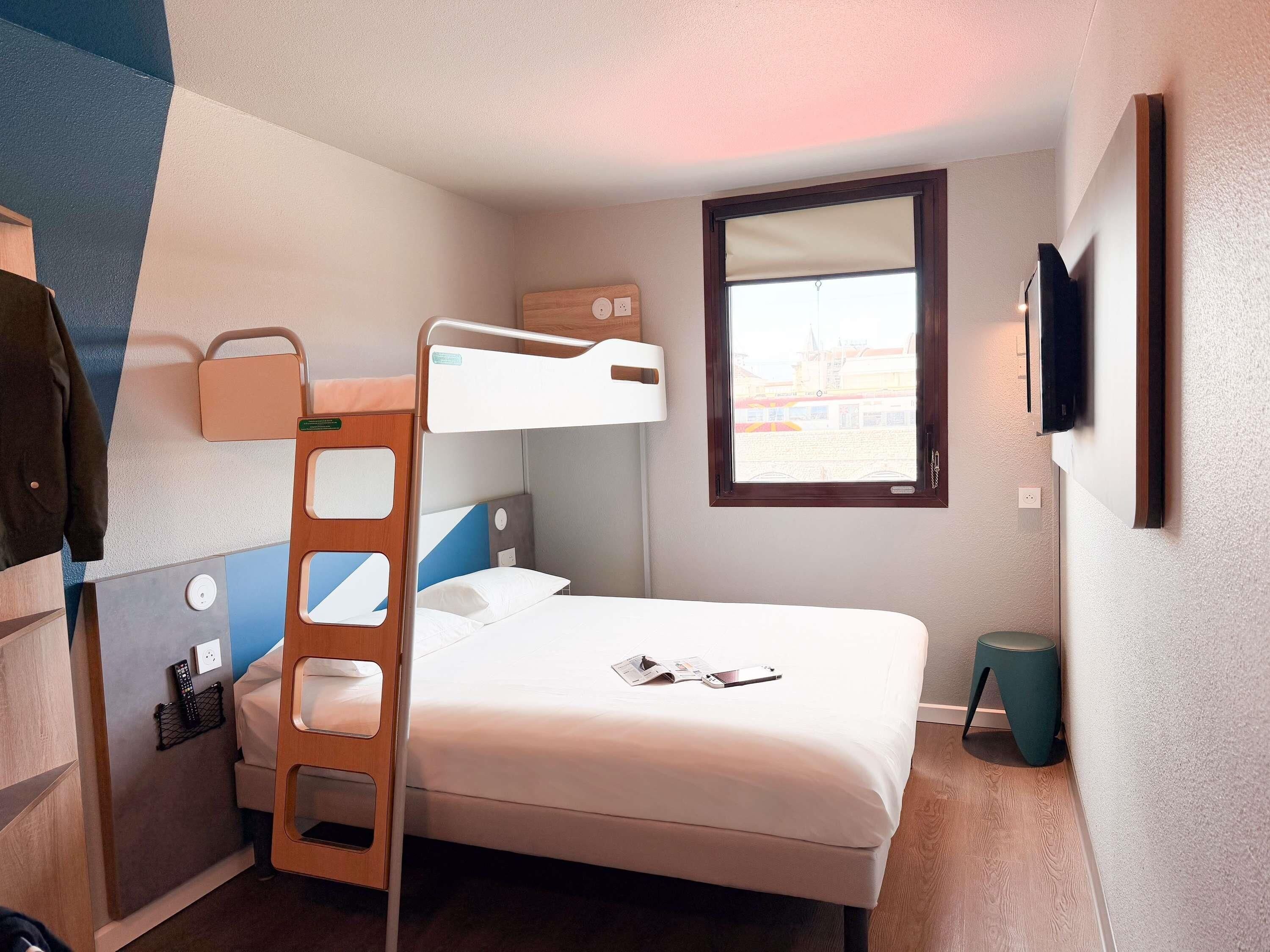 Habitación de hotel compacta con literas, un escritorio pequeño con silla, una mesita de noche, un televisor montado en la pared y una ventana con vista a edificios.