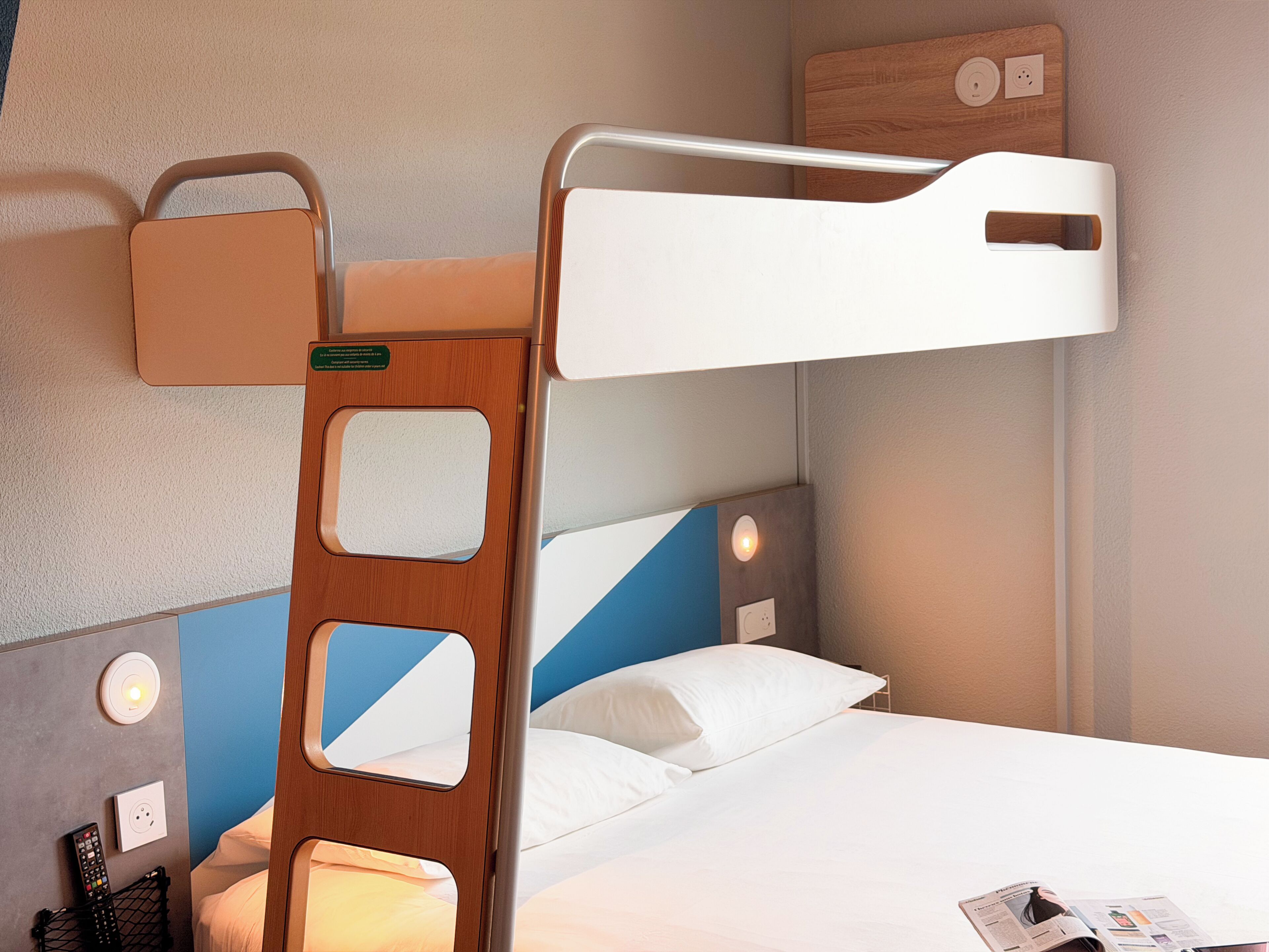 A bunk bed with a ladder, a bedside table, a wall socket, and a light switch.