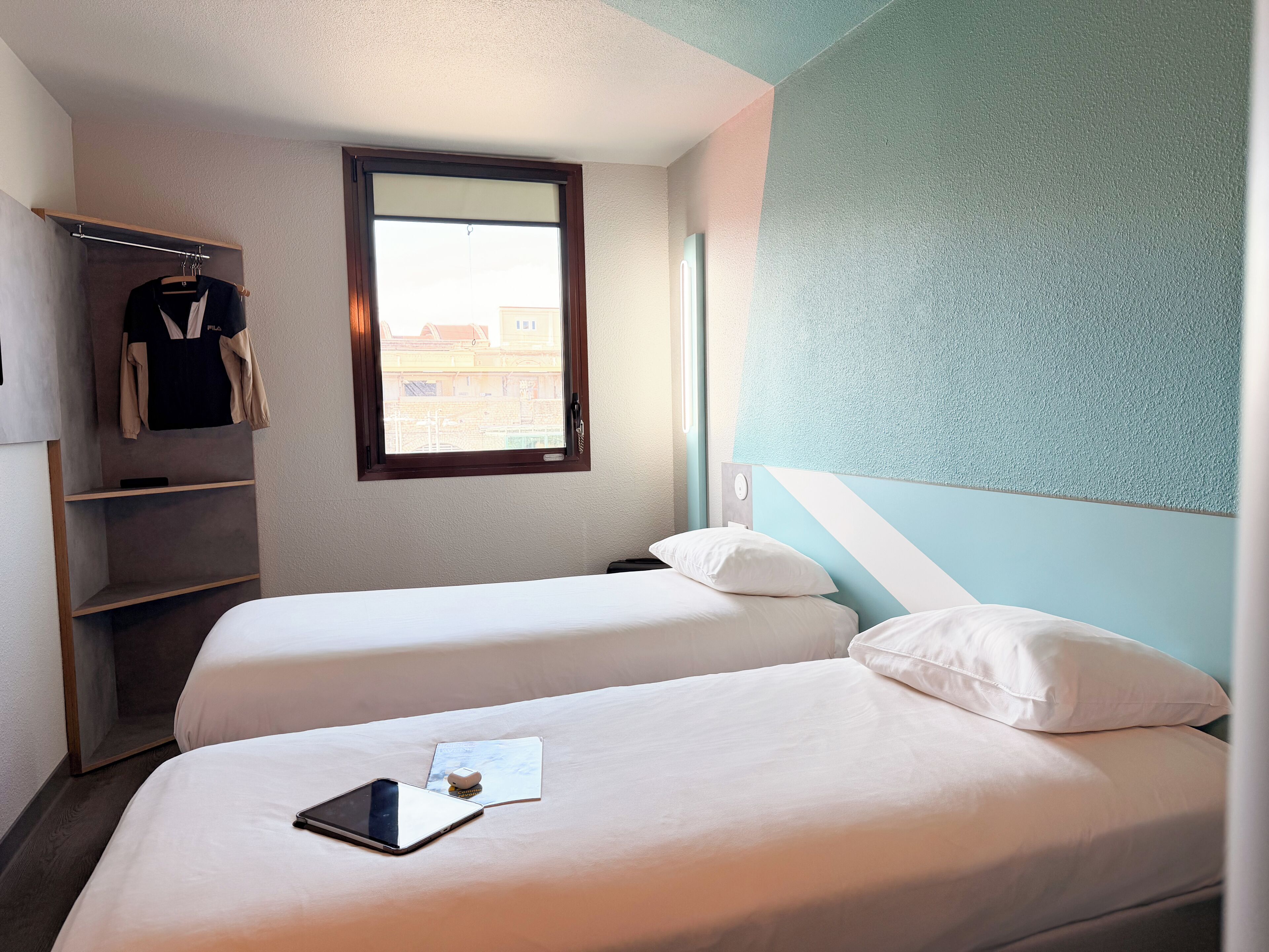 A hotel room with two beds, a window, a coat rack, and a small table with a tablet.