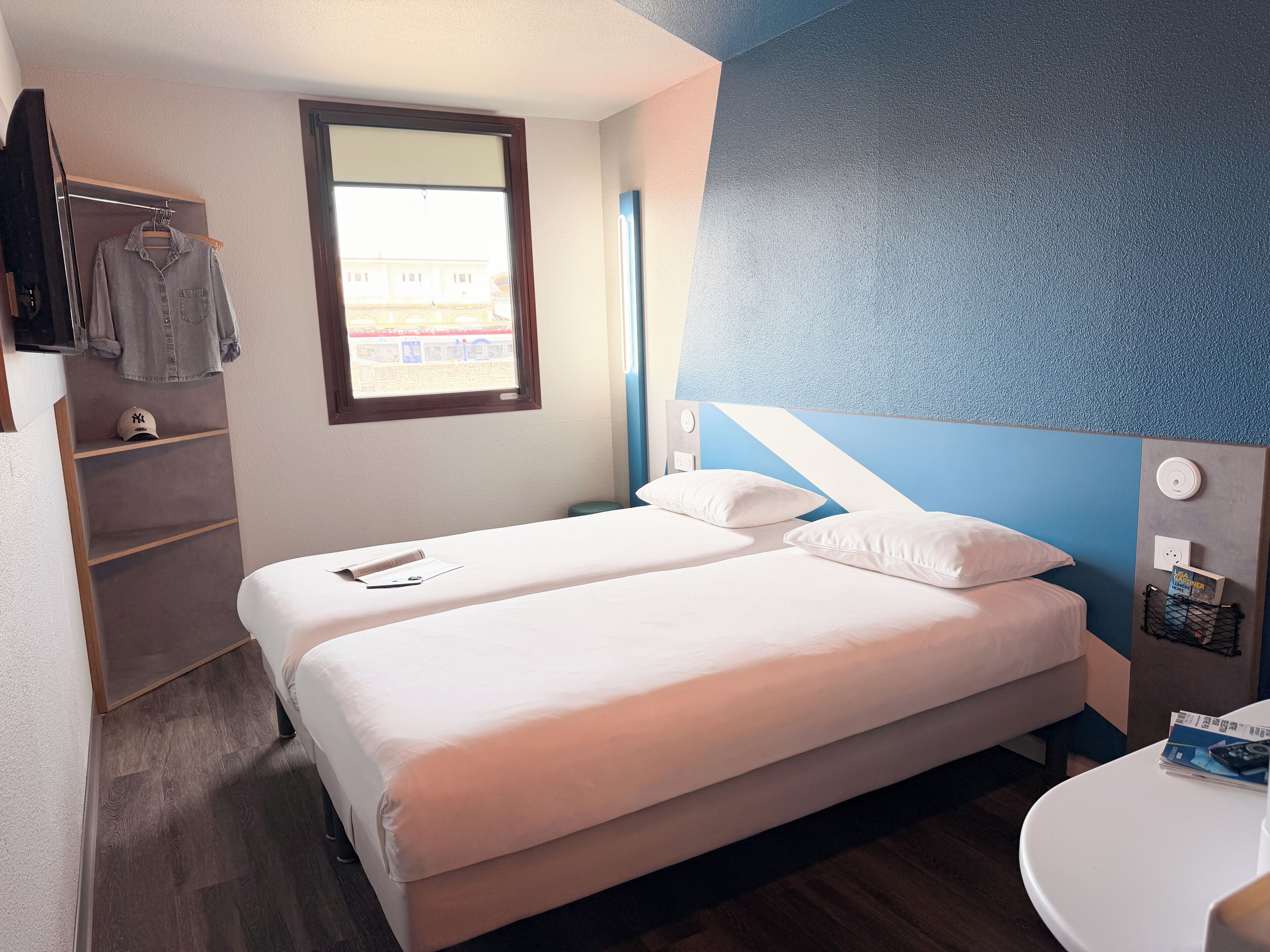 Ein Hotelzimmer mit zwei Betten, einem Fenster, einer blauen Akzentwand, einem Nachttisch mit einem Buch und einem Badezimmer mit einem weißen Waschbecken.