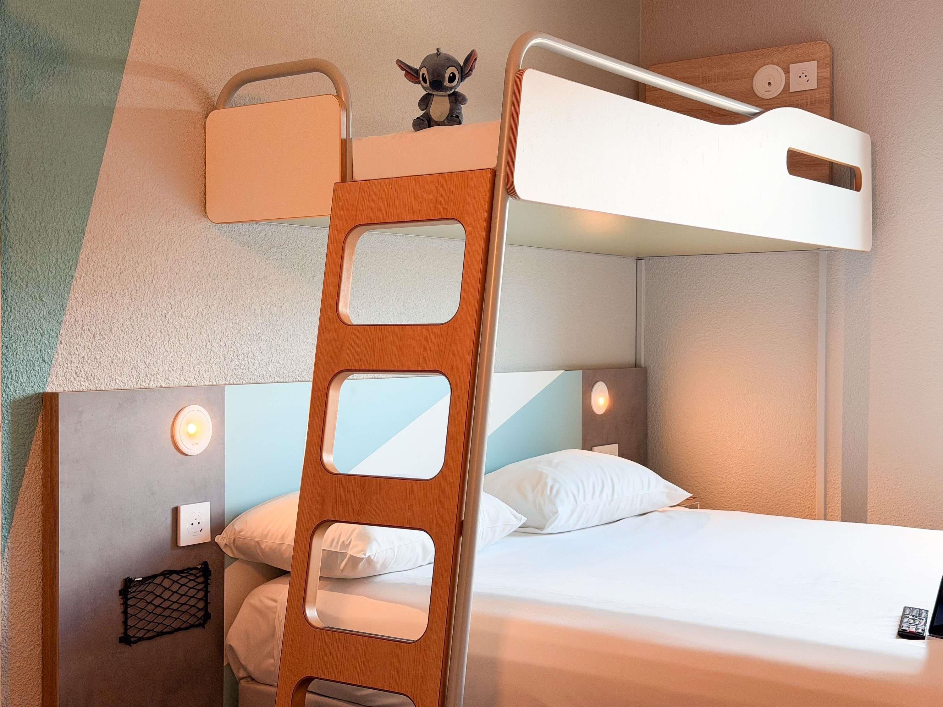 Una cama litera con escalera, un peluche pequeño, lámparas de noche y un control remoto en la cama inferior.