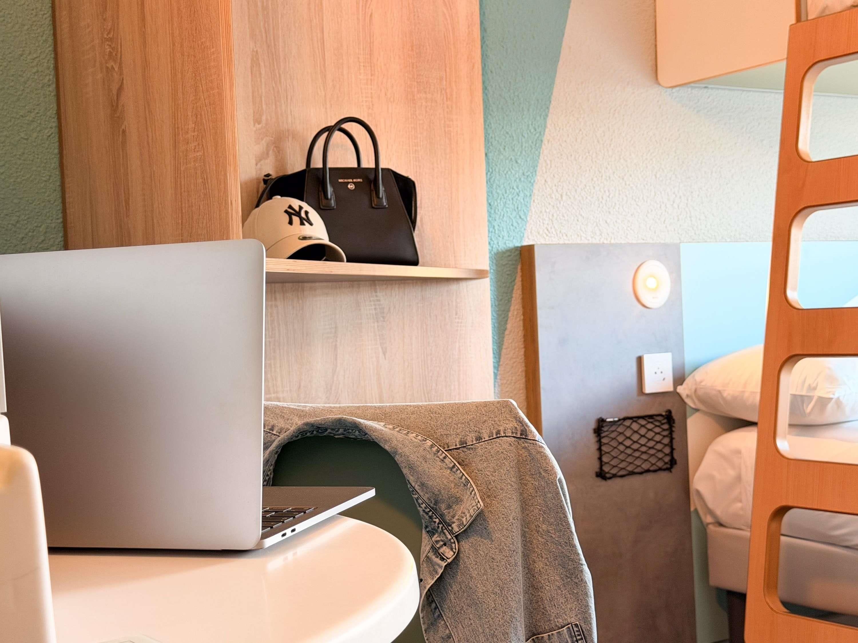 Una habitación de hotel con una laptop sobre una mesa, una cama con ropa blanca, un cabecero de madera y una estantería con un bolso negro.