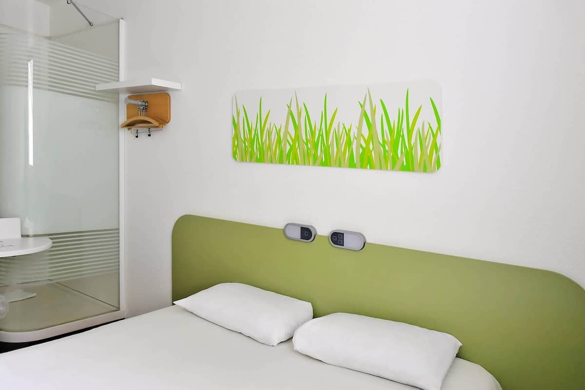Une chambre d’hôtel avec un lit, des oreillers blancs, une tête de lit verte, un panneau de commande fixé au mur et une salle de bain dotée d’une douche à parois de verre.