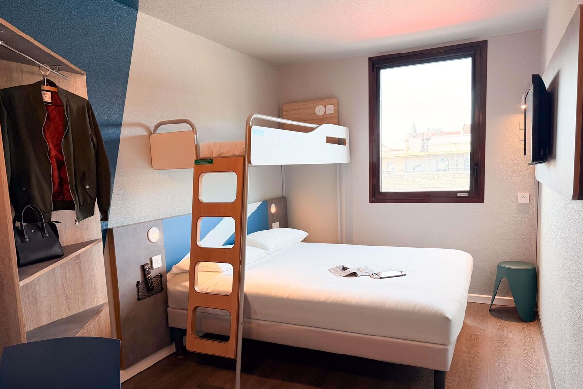 Een stapelbed met een ladder, een bureau met stoel, een hangende kapstok en een raam met uitzicht.