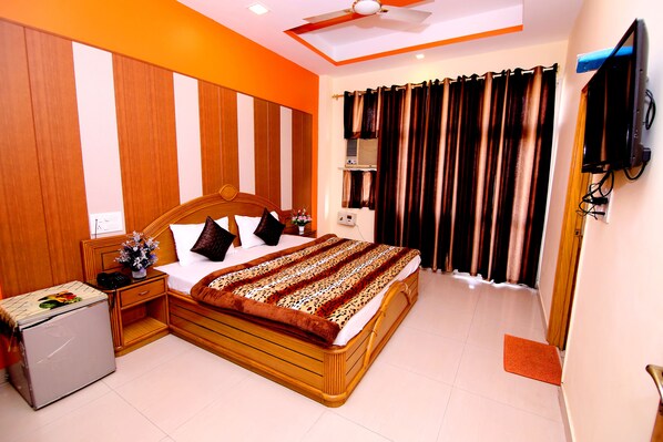 A bedroom with a wooden bed, a small refrigerator, a ceiling fan, and a television mounted on the wall. - Hotel Sunshine (Haridwar)