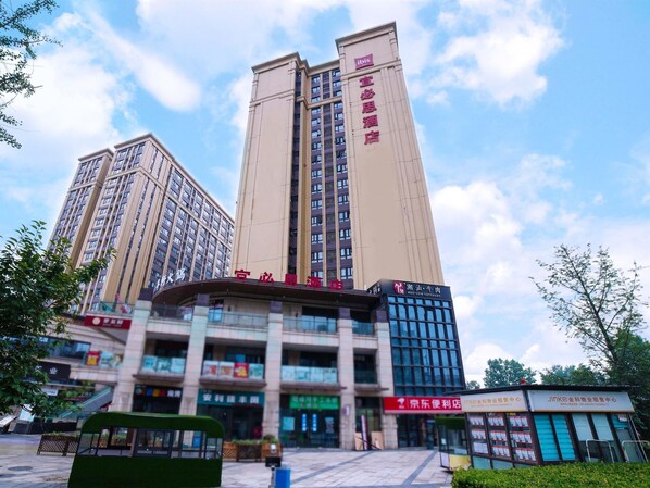 A multi-story hotel with a prominent sign, a bus stop, and surrounding greenery. - ibis Chengdu West Jingcui Road (Chengdu)
