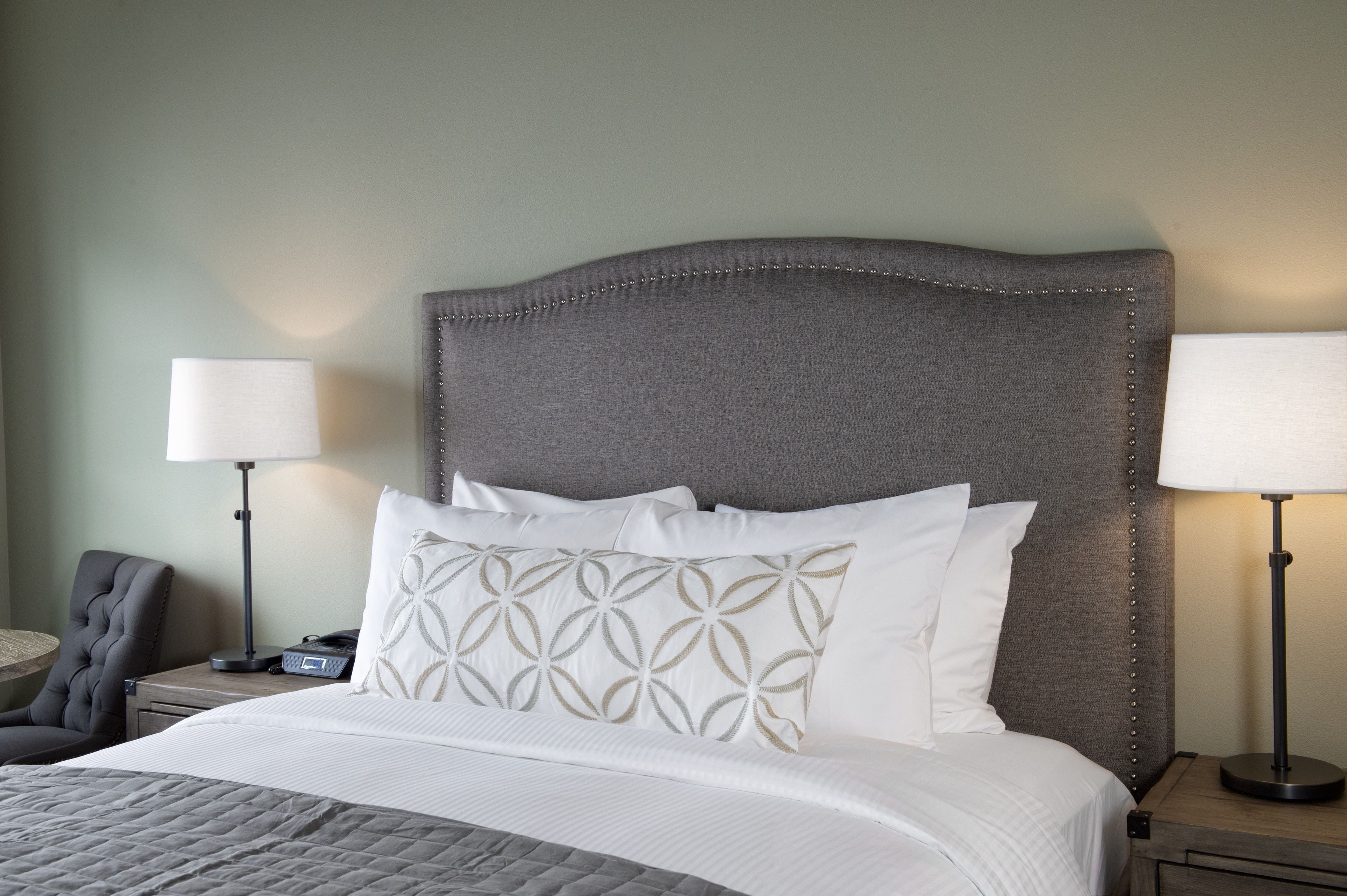 A neatly made bed with white linens and decorative pillows, a gray upholstered headboard, and two bedside lamps on wooden nightstands.