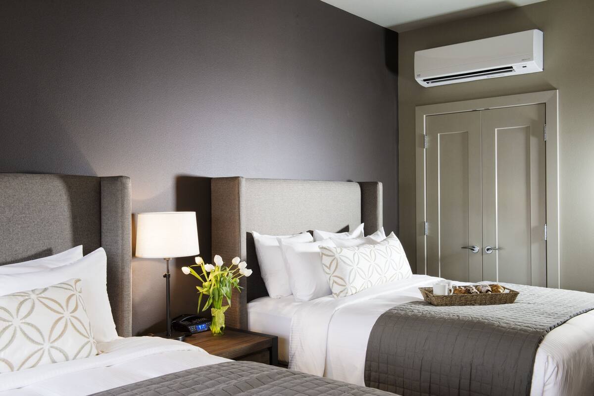 A hotel room with two beds, a nightstand with a lamp, a vase of flowers, and a basket of pastries.