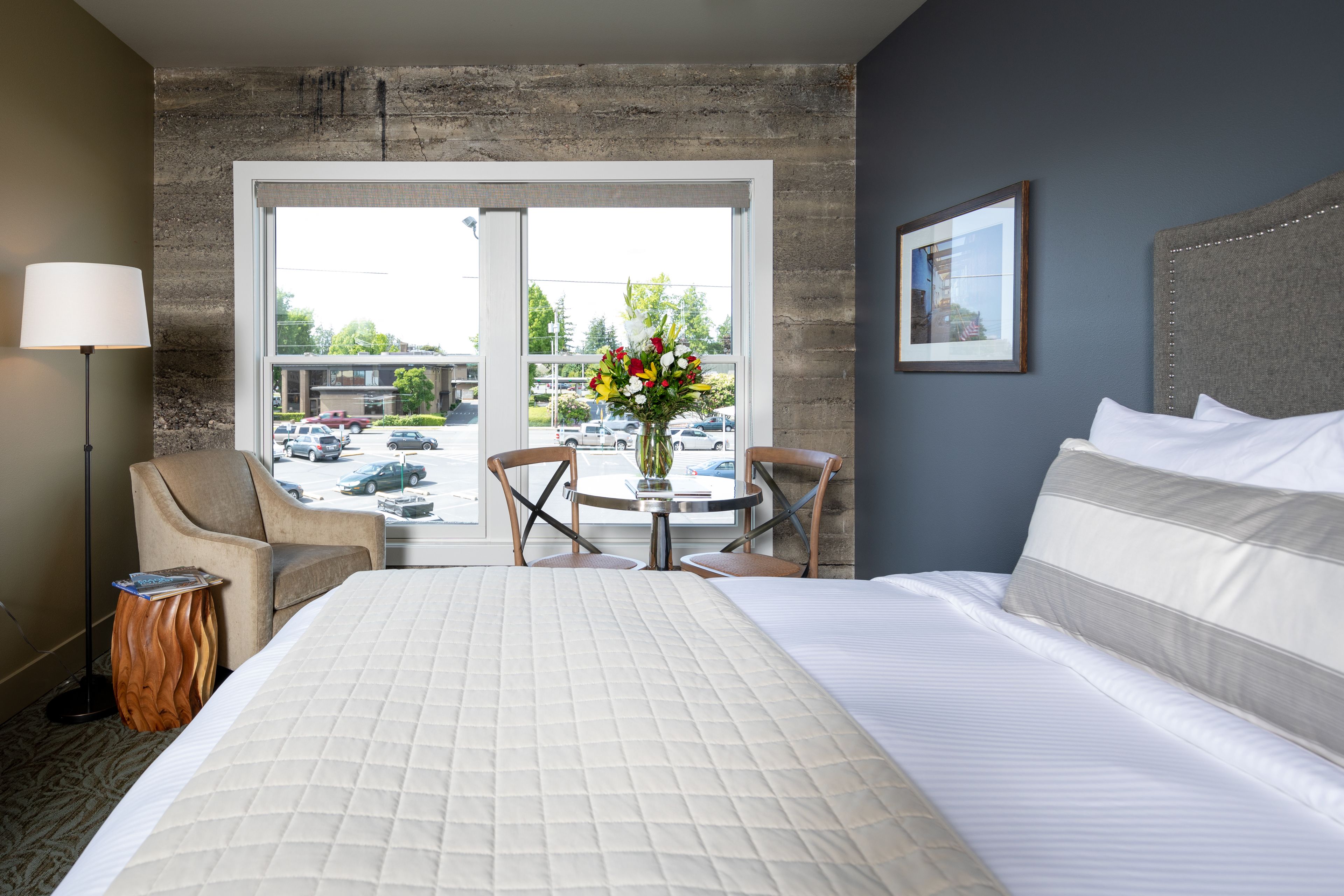A hotel room with a large bed, a desk with chairs, a lamp, a vase with flowers, and a view of the parking lot and buildings outside.