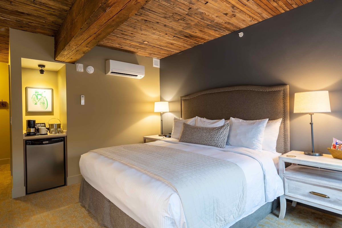 A hotel room with a large bed, two bedside tables with lamps, a small desk with a lamp, an air conditioning unit, and a wall-mounted air purifier.
