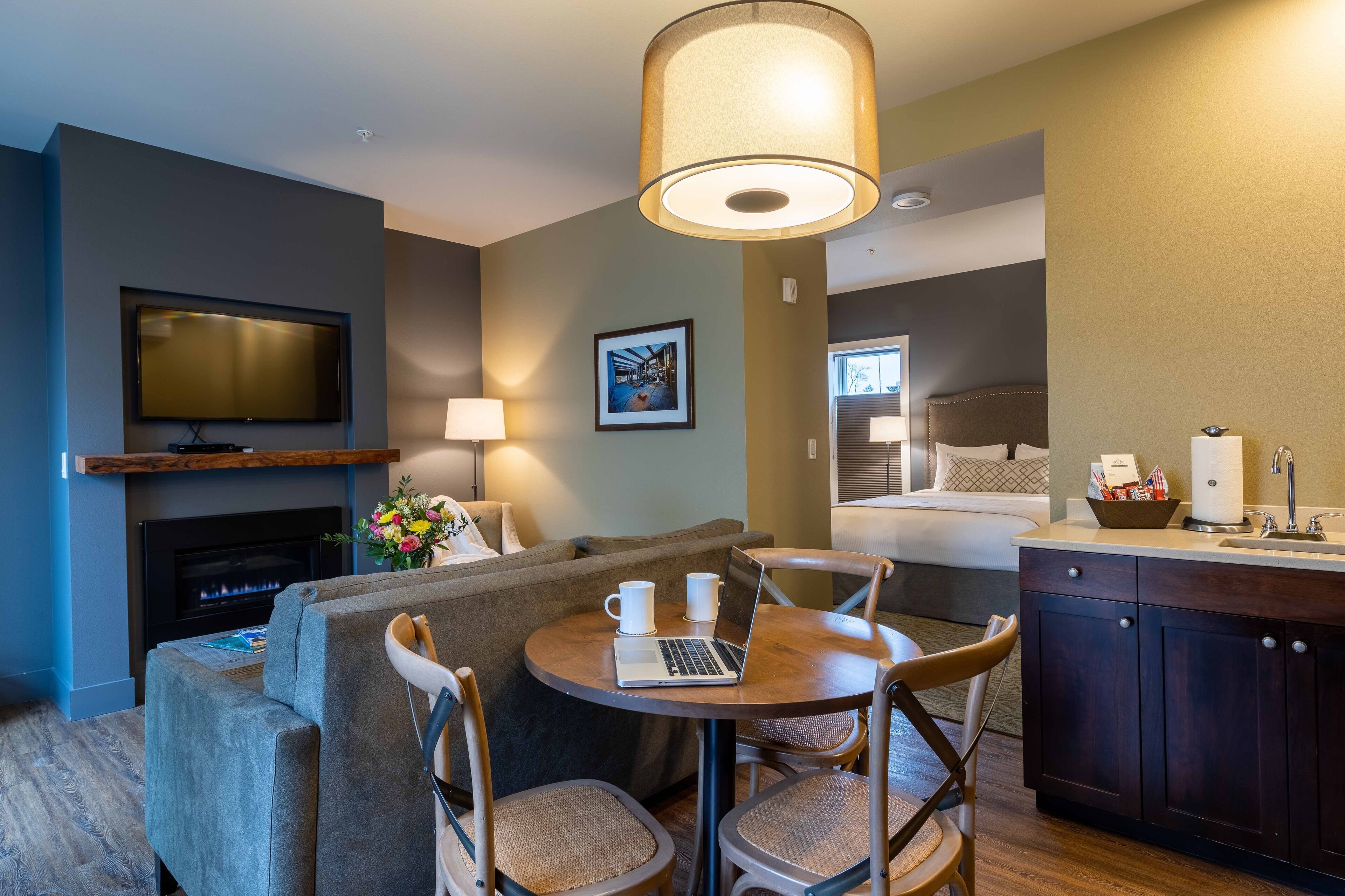 A modern hotel room with a flat-screen TV, fireplace, dining area with a laptop, and a kitchenette.