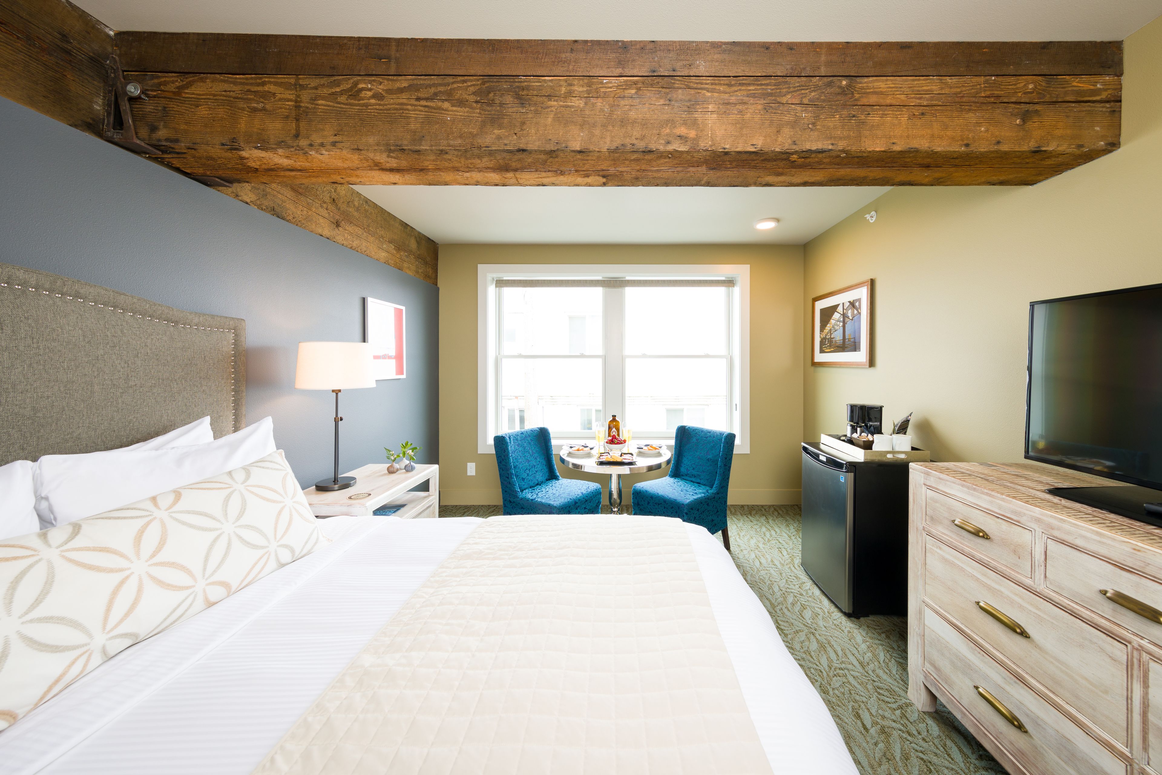 A hotel room with a large bed, a desk with a lamp, two chairs, a television, and a dresser.