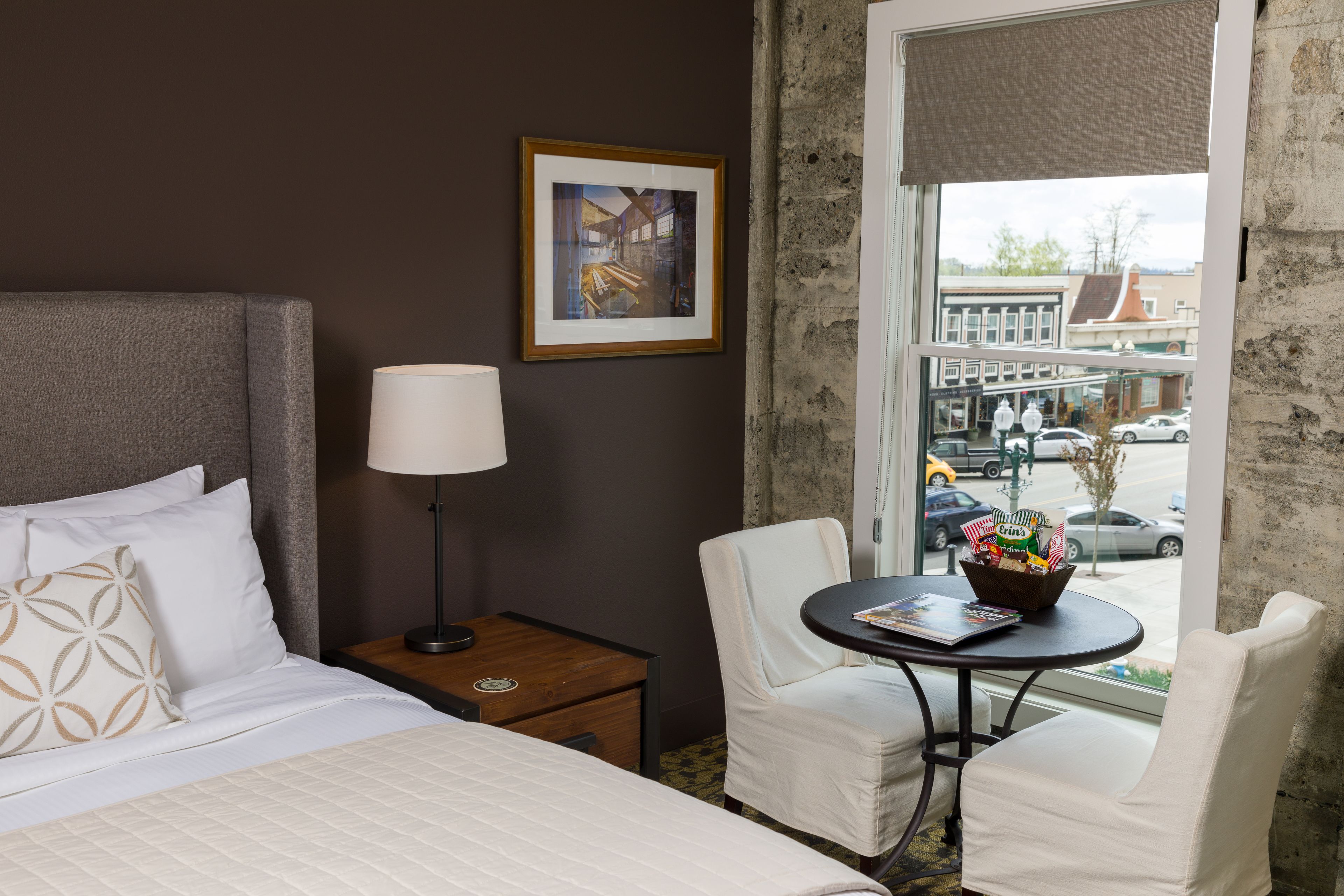 Une chambre d’hôtel avec un lit, une table de chevet, une lampe, une chaise, une table avec un magazine et une fenêtre donnant sur la rue.