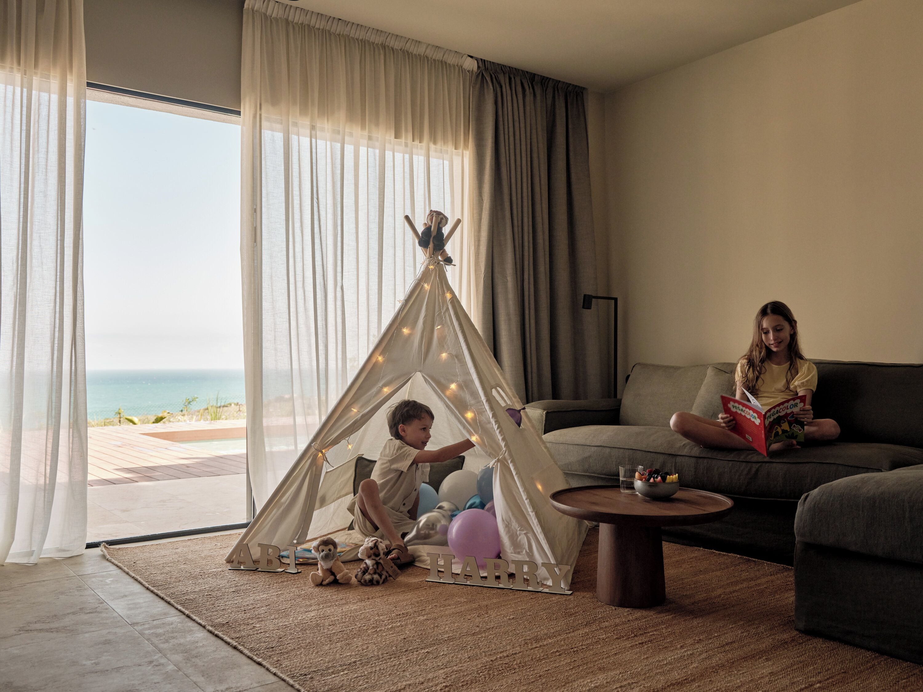 A room with a large window, a sofa, a coffee table, a teepee, and a child reading a book.