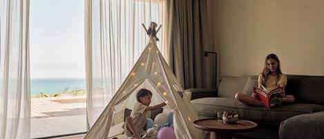 A room with a large window, a sofa, a coffee table, a teepee, and a child reading a book.