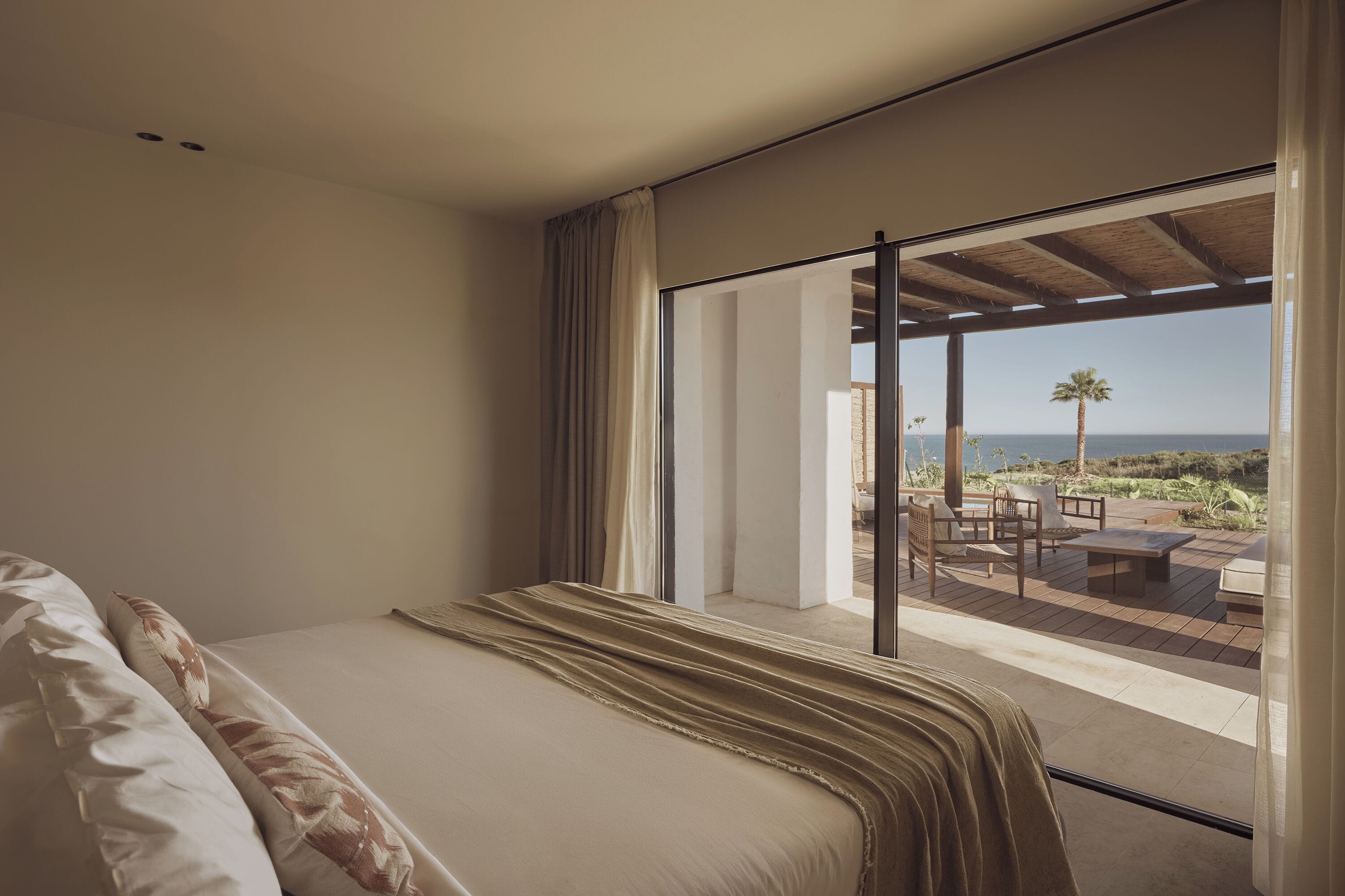 A bedroom with a large bed, a view of an outdoor patio with seating, and a palm tree.