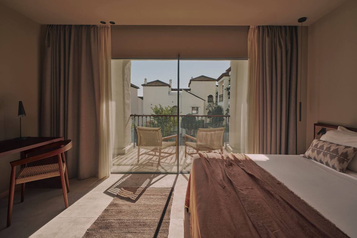 Habitación de hotel con cama, escritorio, silla y balcón con vista a edificios.