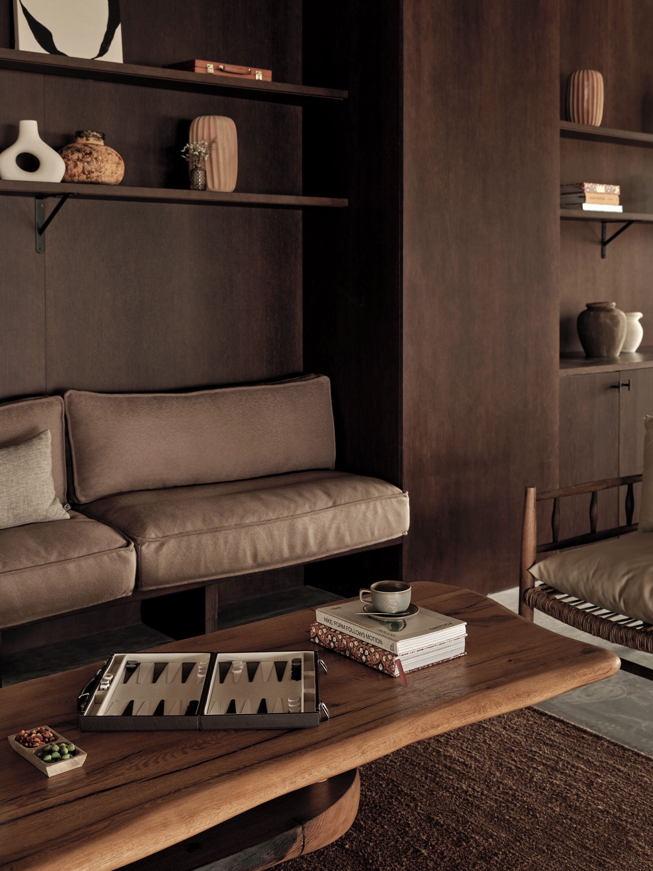 Una sala de estar moderna con un sofá de cuero marrón, una mesa de centro de madera con libros y una bandeja, y estantes con objetos decorativos.