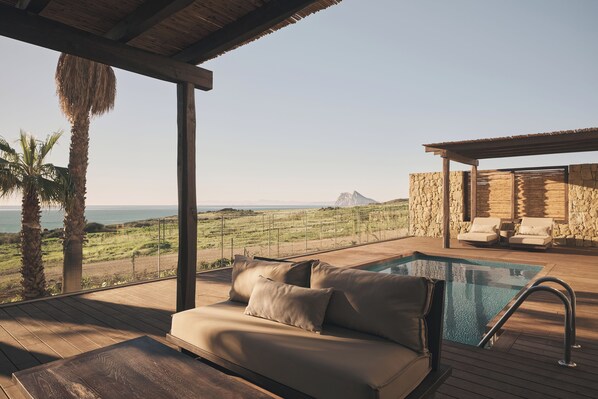 A wooden deck with a pool, a sofa with cushions, and a stone wall. - OKU Andalusia – Alcaidesa (La Línea de la Concepción)