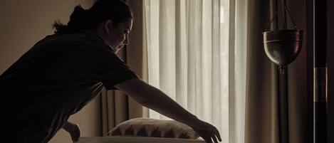 Una persona acomodando una cama en una habitación con poca luz, una ventana y cortinas.