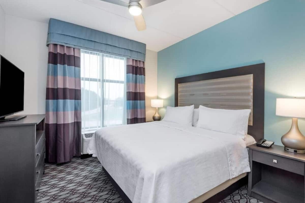 Una habitación de hotel con cama, televisión, escritorio con lámpara y ventana con cortinas.
