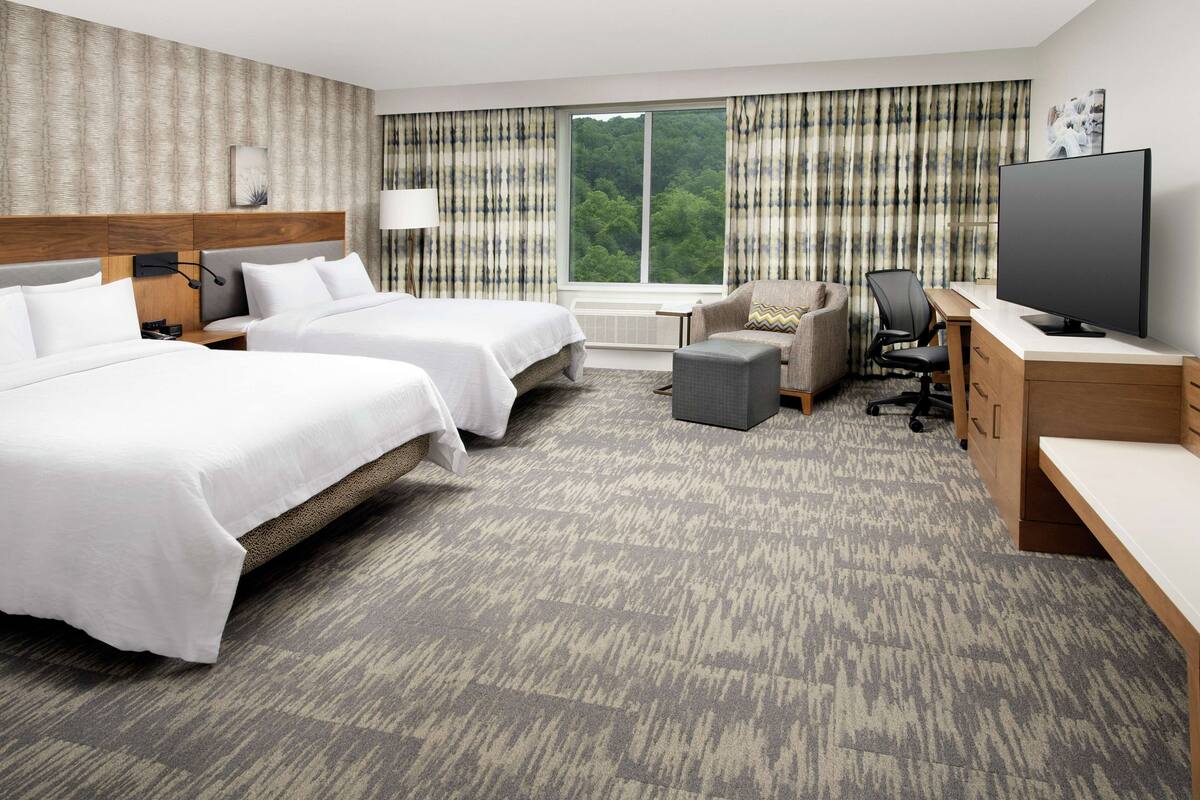 Habitación de hotel con dos camas, un escritorio con televisor, una silla y vistas a un bosque a través de amplios ventanales.