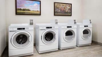 A row of four white front-loading washing machines in a laundry room with two framed landscape paintings on the wall.
