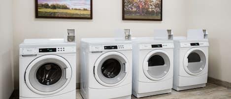 A row of four white front-loading washing machines in a laundry room with two framed landscape paintings on the wall.
