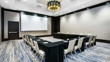 A conference room with a large table, white chairs, a projector screen, and a carpeted floor.