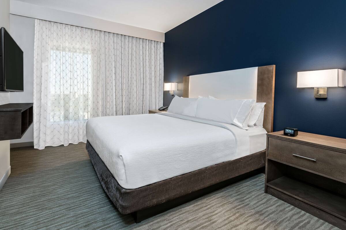 Une chambre d’hôtel avec un grand lit, une table de chevet, un bureau, une télévision et une fenêtre avec des rideaux.