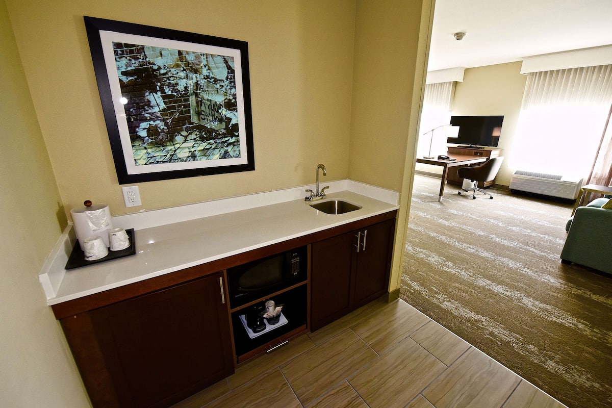 A hotel room with a kitchenette, a flat-screen TV, and a large window with curtains.