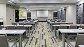 A conference room with rows of tables and chairs, a projector screen, and a carpeted floor.