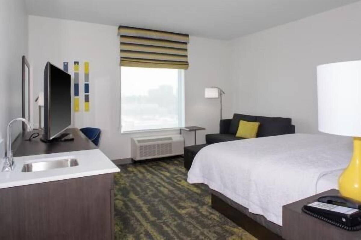 A hotel room with a bed, a desk with a computer, a sink, a window with blinds, and a lamp.