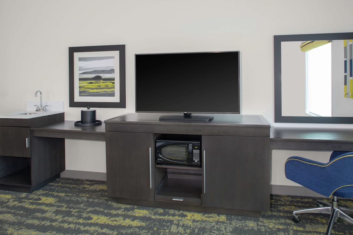 A hotel room with a desk, a flat-screen TV, a microwave, a sink, and a chair.