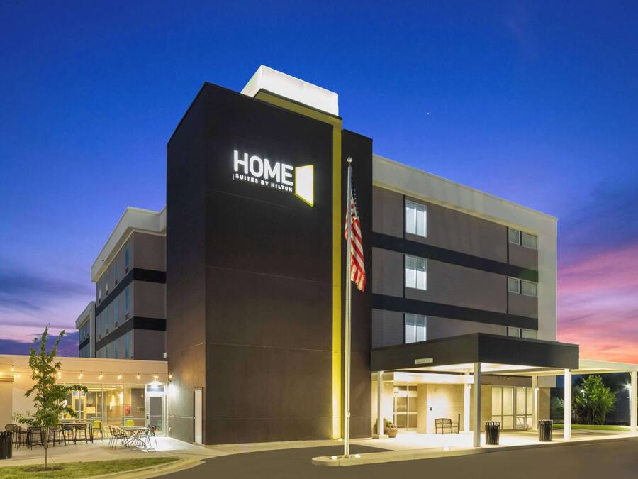A modern hotel building with a "HOME Suites by Hilton" sign, a flag, and a covered outdoor seating area.
