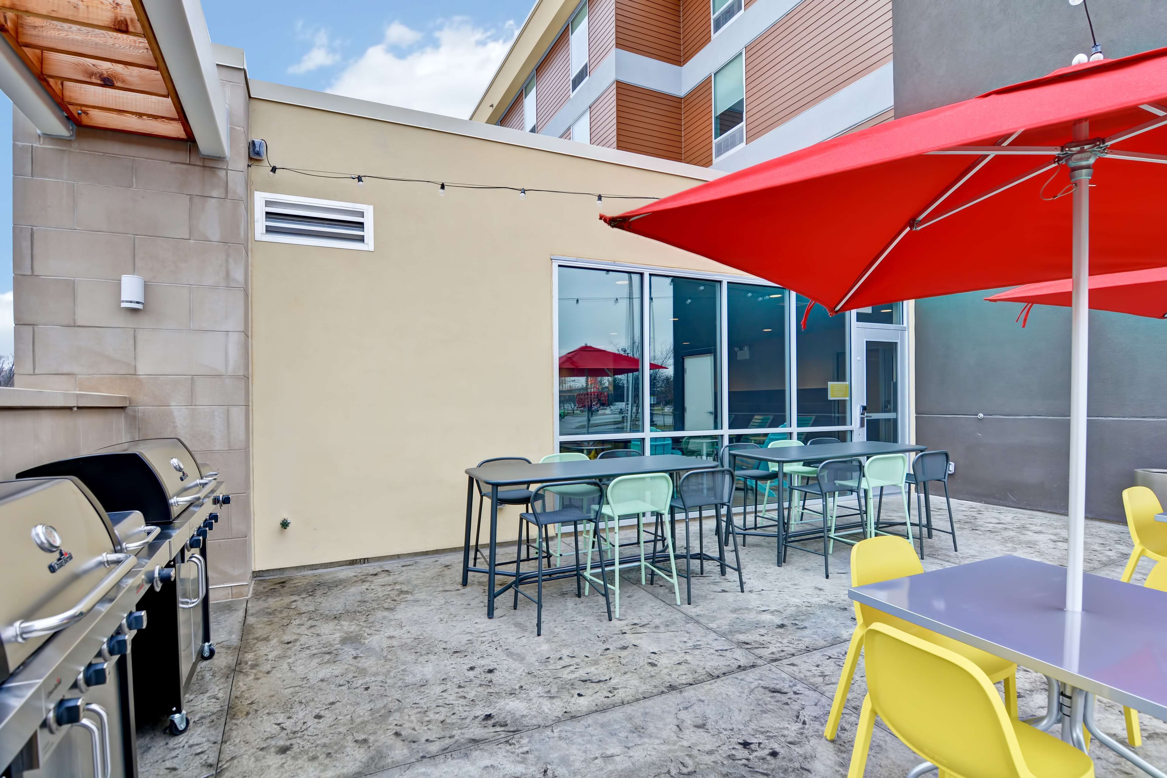 Un patio al aire libre con una parrilla, mesas, sillas y un paraguas rojo.