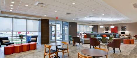 A spacious indoor area with a mix of seating options including tables, chairs, and sofas. There are large windows providing natural light and views of the outside.