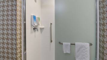 Un área de ducha con mampara de vidrio, paredes de azulejos blancos, un pasamanos y un perchero con dos toallas blancas.